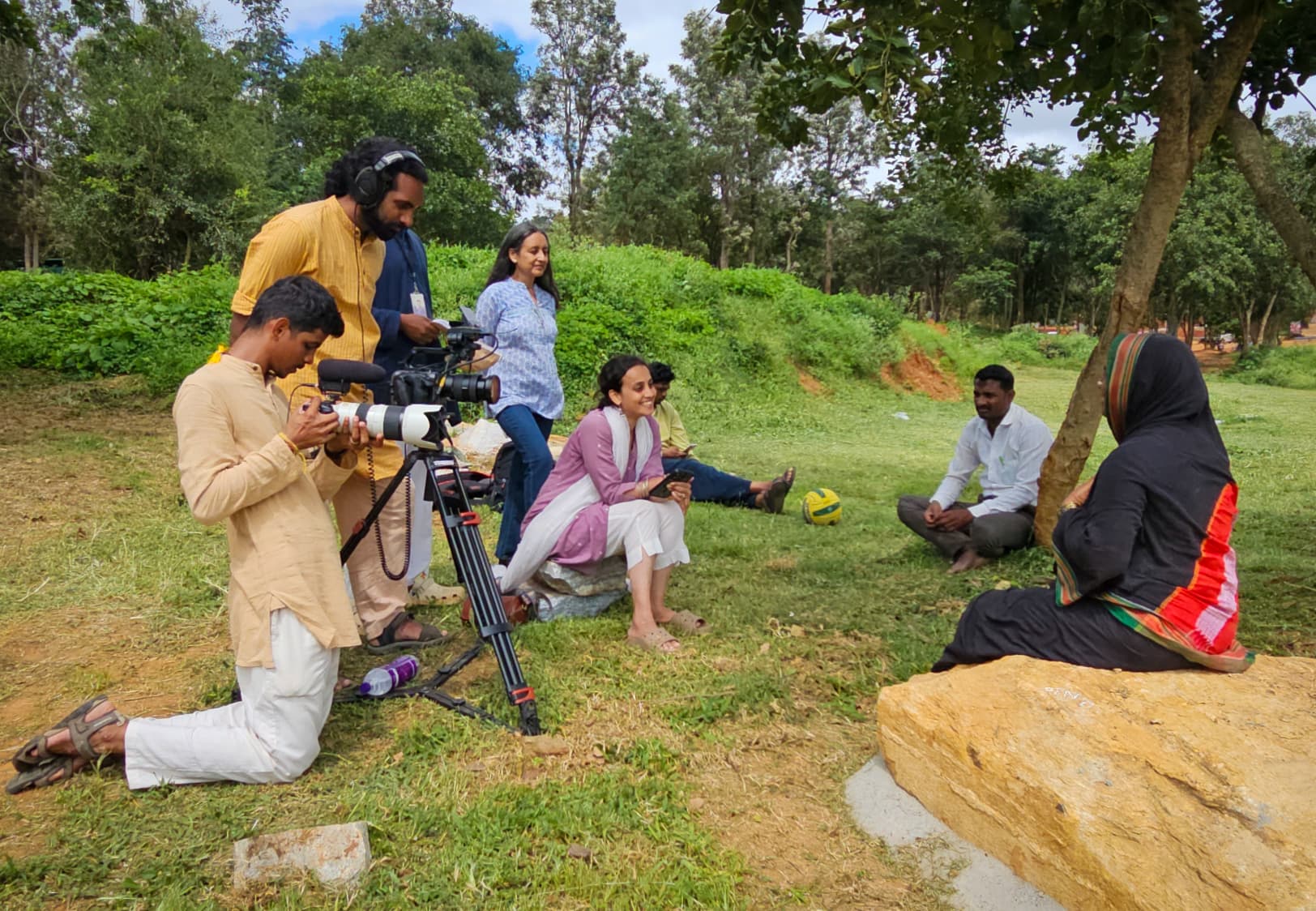 Camera crew filming villagers seated under a tree on location